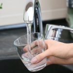 Hand holding a glass under a faucet with clean water flowing, emphasizing safe drinking water and backflow prevention importance.